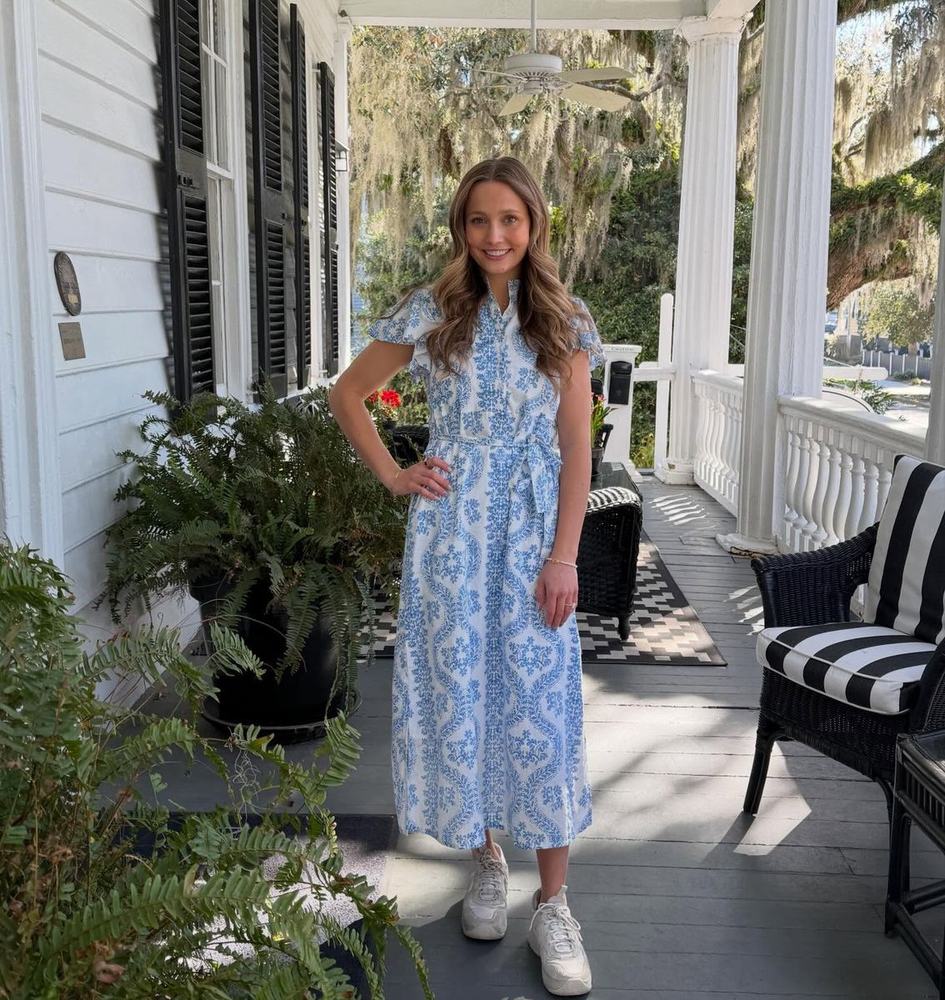 Isabelle, the maker behind Stitched by Izz, standing on a porch in a blue-and-white dress