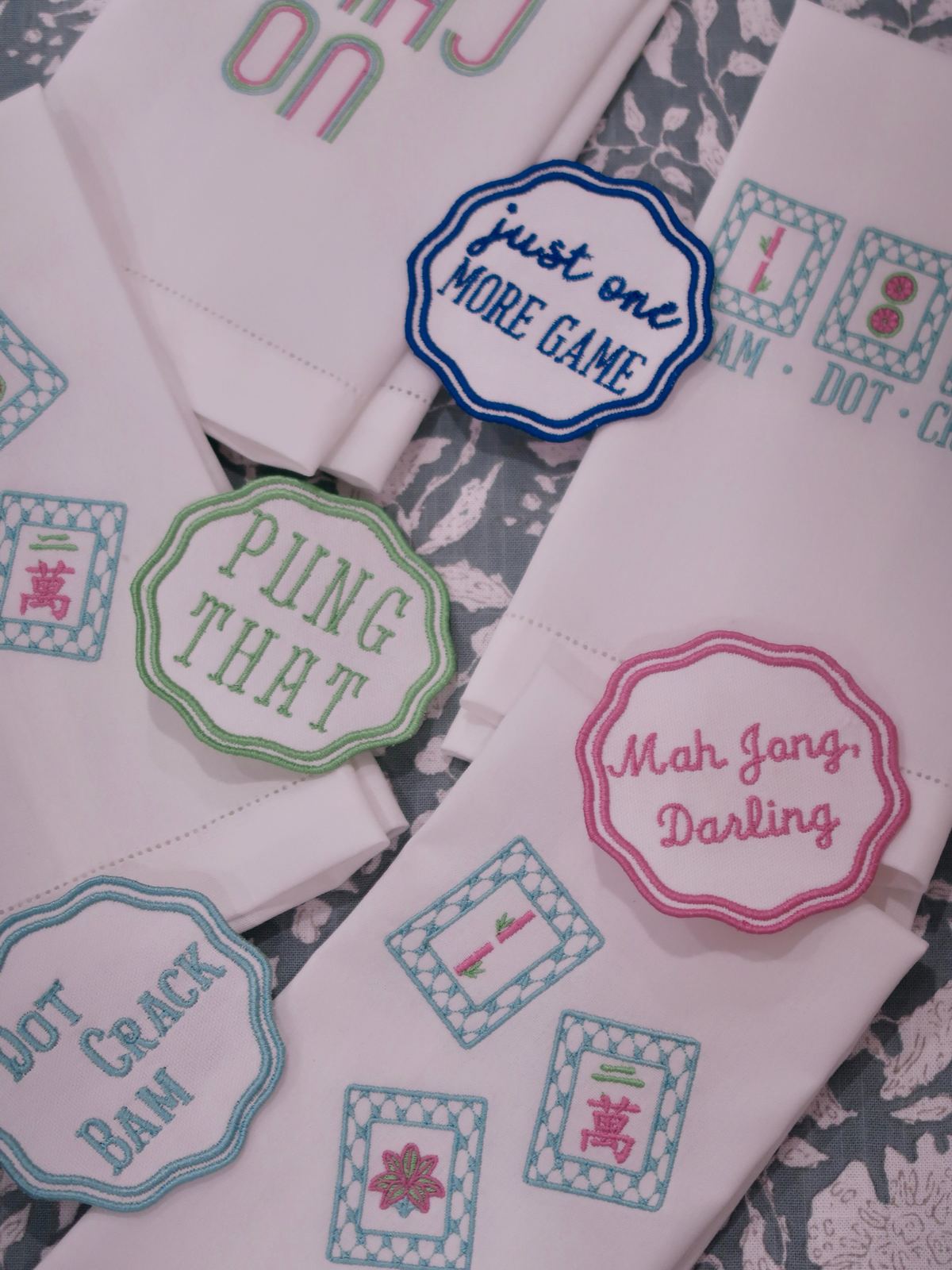 Embroidered mahjong coasters reading Just one more game, Pung that, Mah Jong Darling and Dot Crack Bam on white hand towels